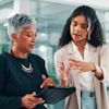 Two professional women discussing business strategy in a modern glass office environment
