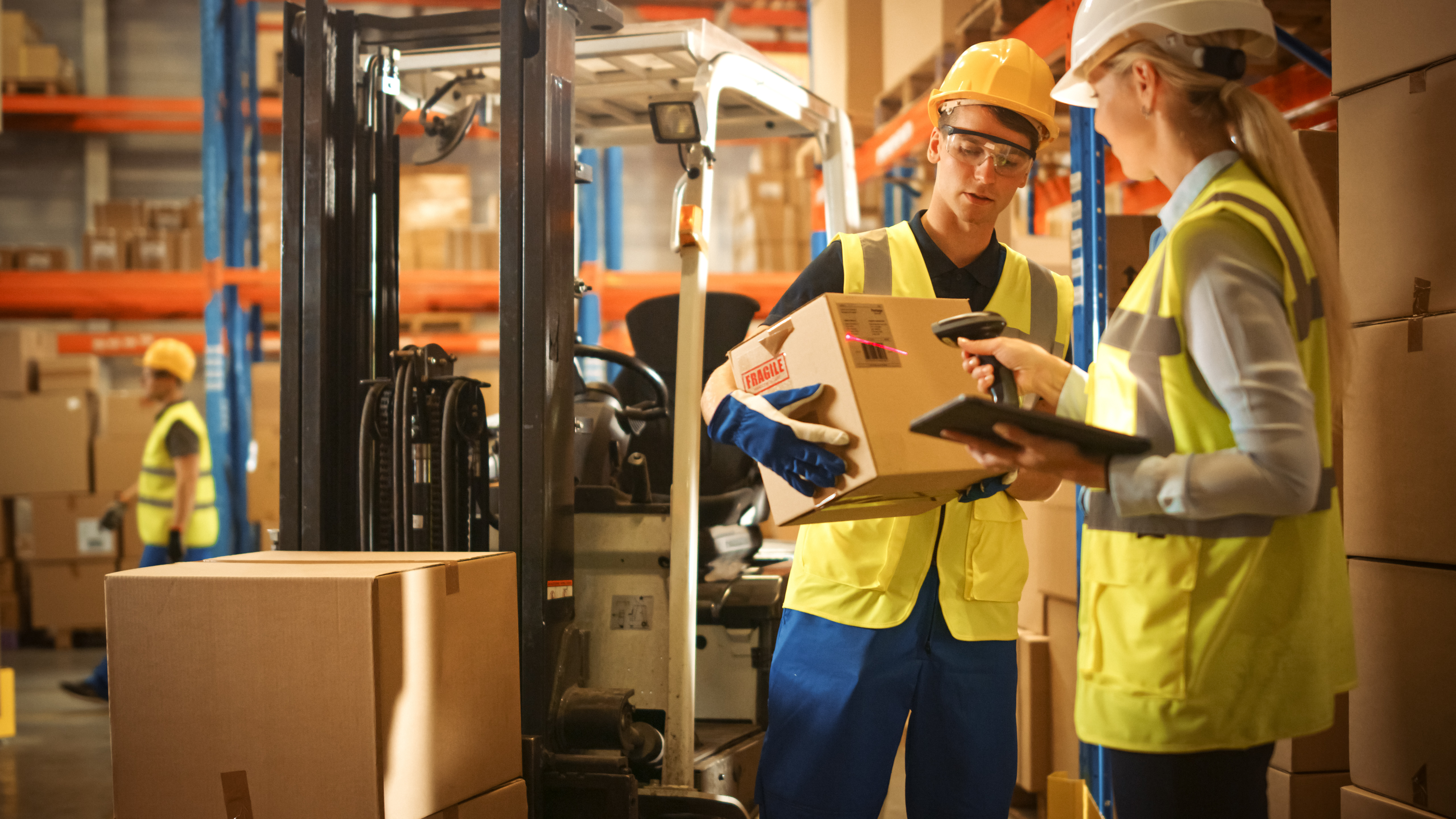 Warehouse workers in safety gear inspect a cardboard box near a forklift in a logistics facility.