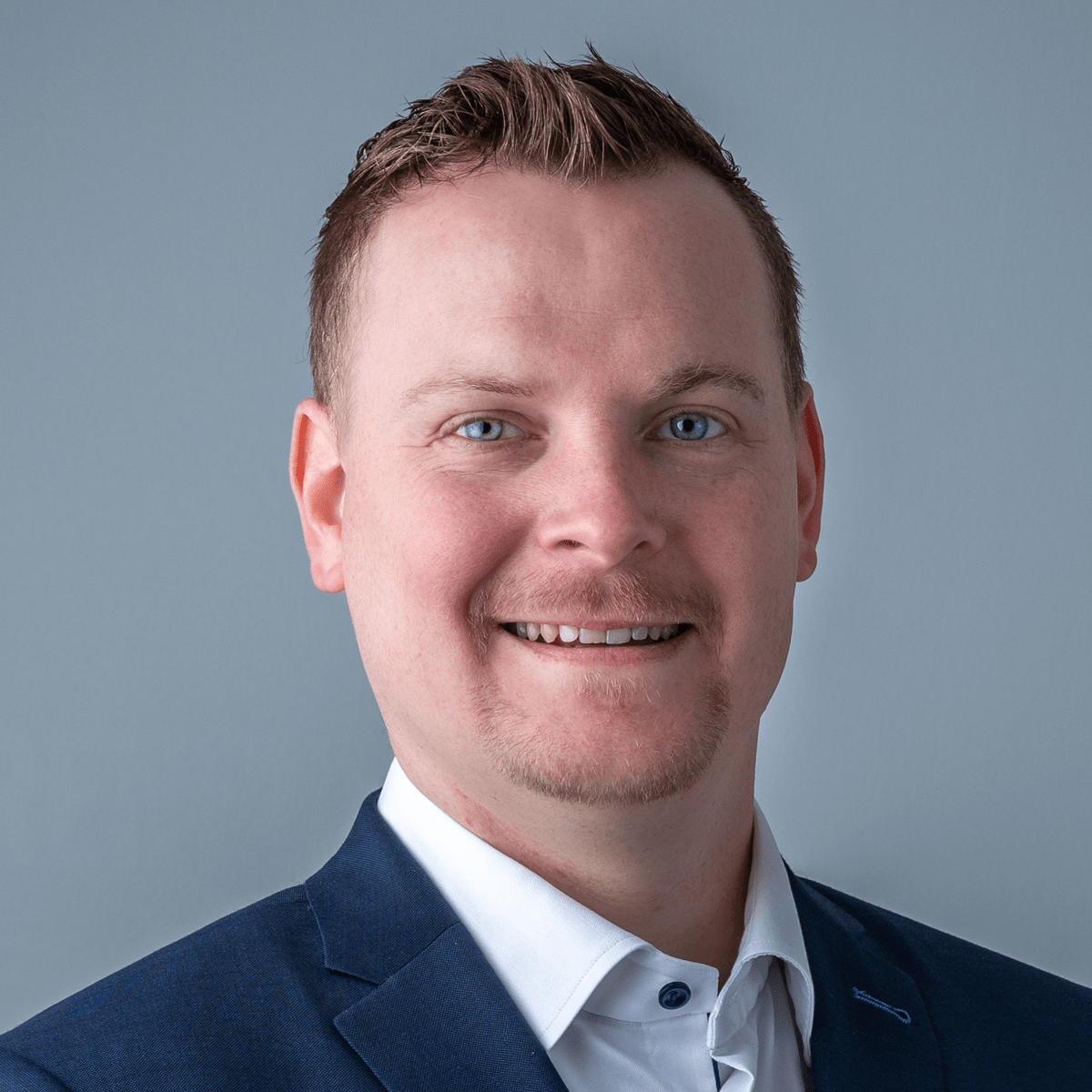 Professional headshot of a smiling man in dark blazer and white shirt against blue background