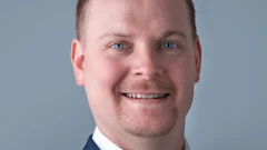 Professional headshot of a smiling man in dark blazer and white shirt against blue background