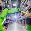 Green robotic arm with yellow gripper operating in a modern warehouse aisle lined with storage shelves.