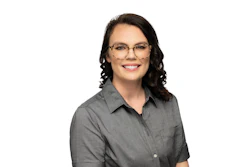 Professional headshot of a woman wearing glasses and a gray button-up shirt, smiling at camera
