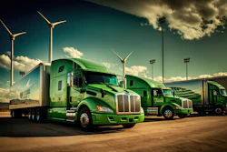 Fleet of green semi-trucks parked at facility with wind turbines and stadium lights under cloudy sky