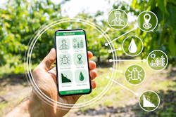 Hand holding smartphone displaying agriculture app with farming icons and crop management tools in field setting