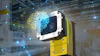 Industrial vision camera with white and black housing mounted on yellow equipment in a smart factory warehouse with digital network visualization
