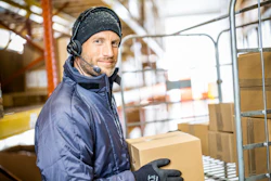 Warehouse worker wearing headphones and winter gear handling cardboard boxes in a logistics facility