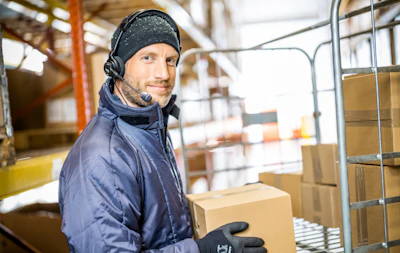 Warehouse worker wearing headphones and winter gear handling cardboard boxes in a logistics facility