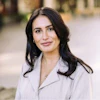 Professional headshot of woman with dark wavy hair wearing light gray blazer