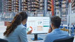 Two professionals reviewing analytics dashboards on a monitor in a warehouse with storage racks in the background