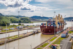 Large cargo ship transiting through Panama Canal locks with container stacks and industrial port infrastructure visible