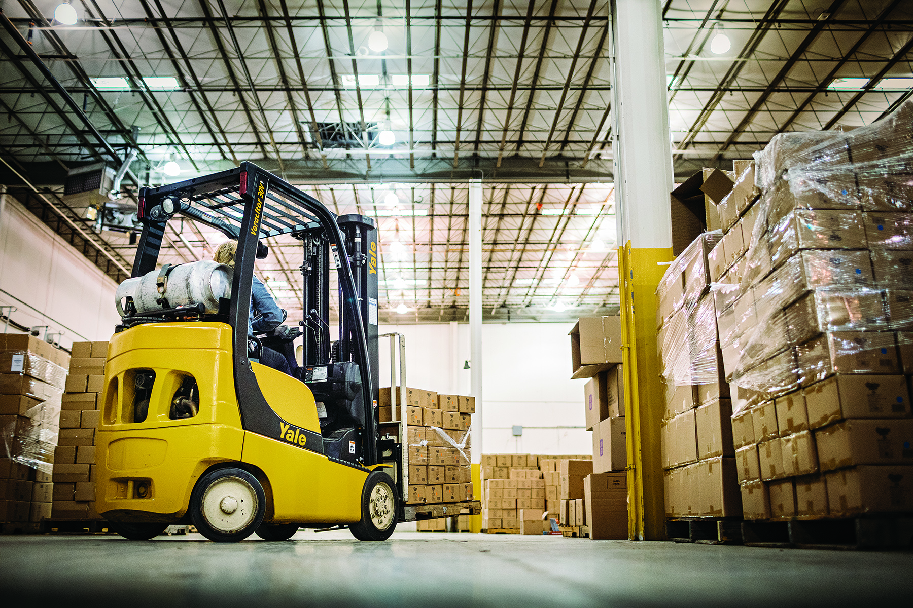 Yellow Yale forklift moving cardboard boxes in a warehouse with stacked pallets and industrial ceiling