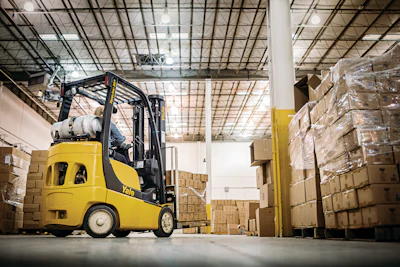 Yellow Yale forklift moving cardboard boxes in a warehouse with stacked pallets and industrial ceiling