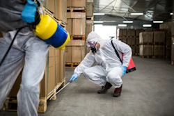 Worker in white protective suit and respirator spraying treatment on warehouse boxes