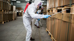 Worker in white protective suit spraying disinfectant on cardboard boxes in warehouse facility