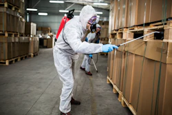 Worker in white protective suit spraying disinfectant on cardboard boxes in warehouse facility