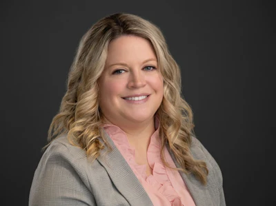 Professional headshot of a woman with blonde wavy hair wearing a beige blazer and pink blouse, smiling against a gray background