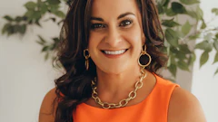 Professional headshot of woman in orange sleeveless top with gold chain necklace, smiling against green plant background