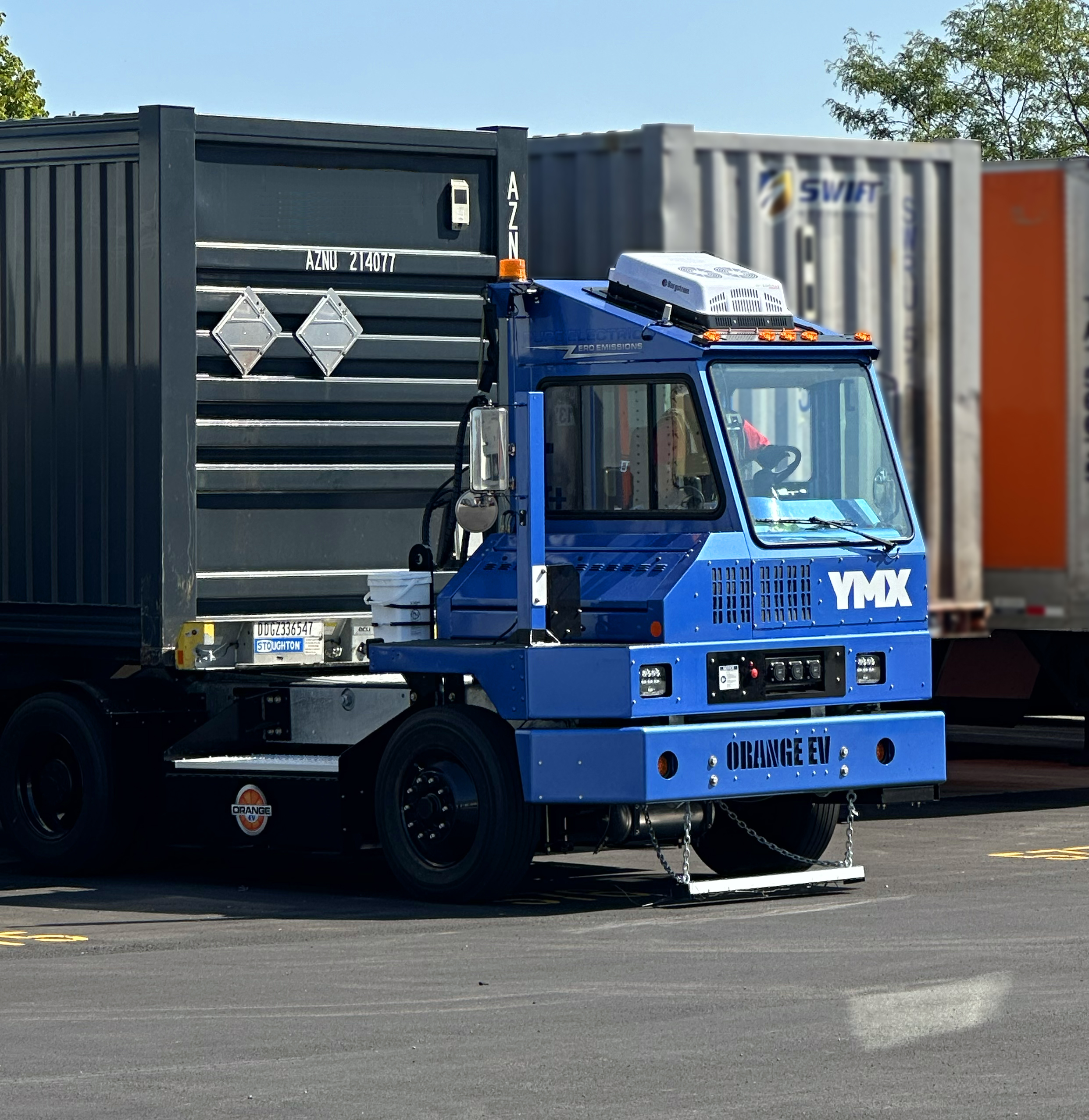 Blue industrial container handling truck with hydraulic crane lifting a dark shipping container in a logistics yard