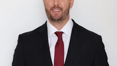 Professional headshot of a man in black blazer and burgundy tie against white background