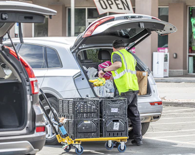 Delivery worker in safety vest loading packages into white van trunk with utility cart in foreground