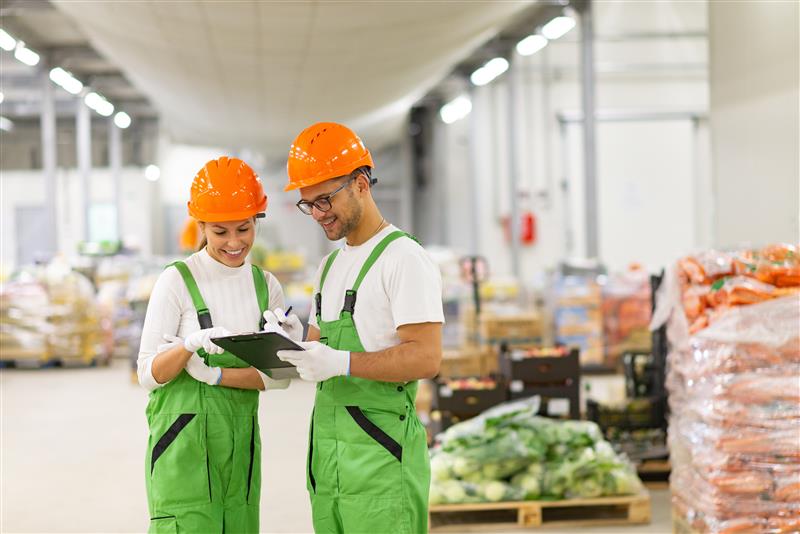 Two smiling warehouse workers in green uniforms and orange hard hats reviewing a clipboard together in a modern industrial facility.