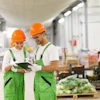 Two smiling warehouse workers in green uniforms and orange hard hats reviewing a clipboard together in a modern industrial facility.