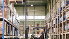 Forklift operator moving cargo in a large warehouse with blue and metal shelving units.