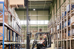 Forklift operator moving cargo in a large warehouse with blue and metal shelving units.