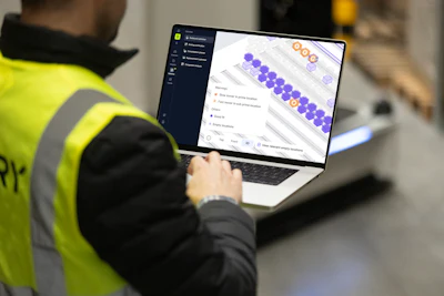 Worker in high-visibility vest using laptop displaying purple and orange data visualization interface in warehouse setting