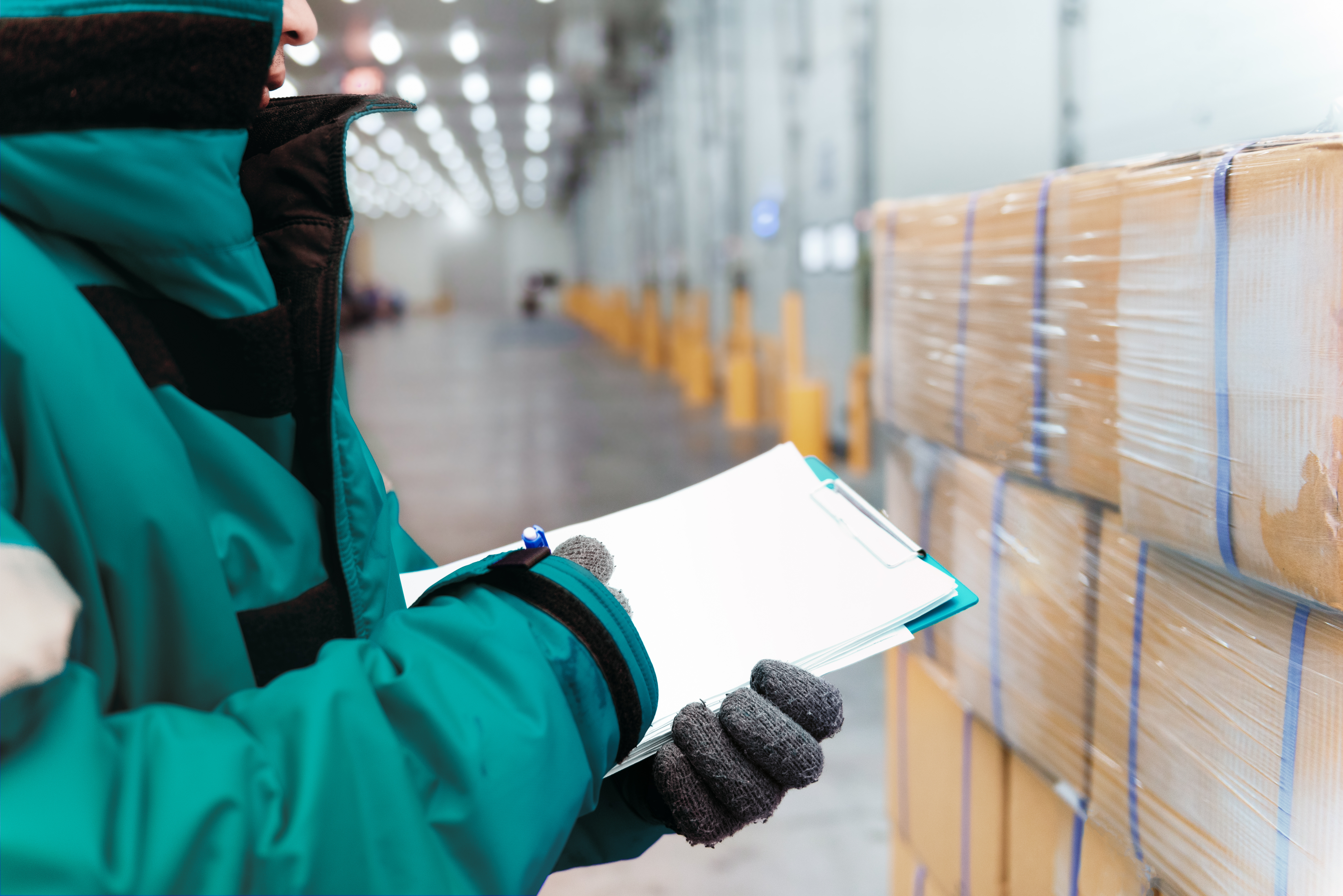 Warehouse worker in teal jacket inspecting stacked cardboard boxes with clipboard