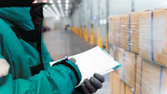 Warehouse worker in teal jacket inspecting stacked cardboard boxes with clipboard