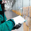 Warehouse worker in teal jacket inspecting stacked cardboard boxes with clipboard