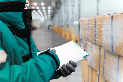 Warehouse worker in teal jacket inspecting stacked cardboard boxes with clipboard