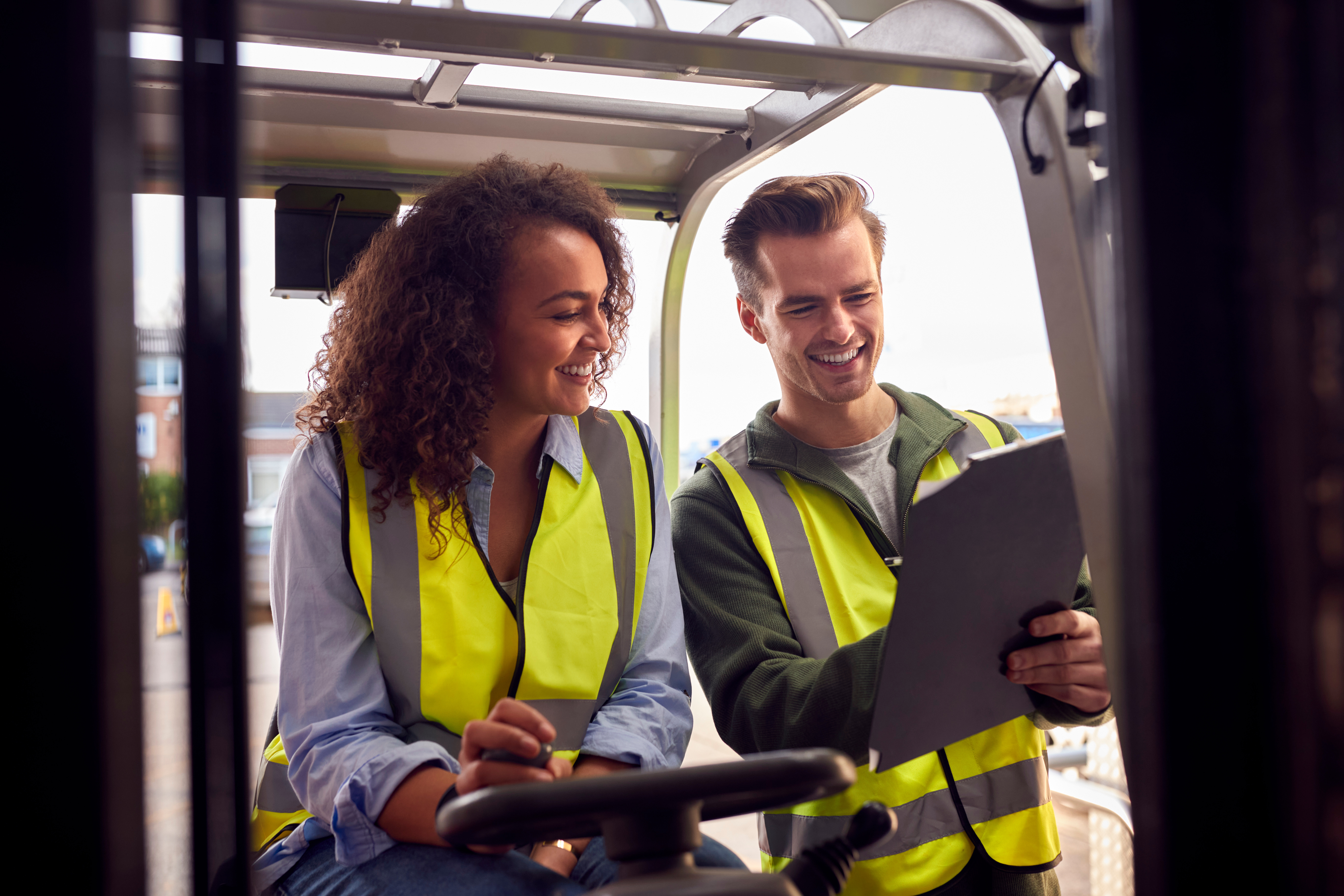 Two smiling workers in yellow safety vests reviewing documents on a tablet outdoors at a construction site