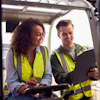 Two smiling workers in yellow safety vests reviewing documents on a tablet outdoors at a construction site
