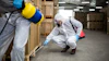 Worker in white protective suit and blue gloves spraying treatment on warehouse shelving with safety equipment visible