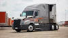 Silver and black Freightliner semi-truck with orange accents parked in shipping yard with cargo containers