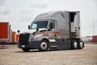 Silver and black Freightliner semi-truck with orange accents parked in shipping yard with cargo containers