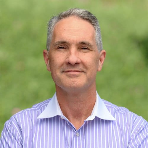 Professional headshot of a middle-aged man wearing a light purple striped shirt with white collar against a blurred green background