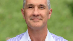 Professional headshot of a middle-aged man wearing a light purple striped shirt with white collar against a blurred green background